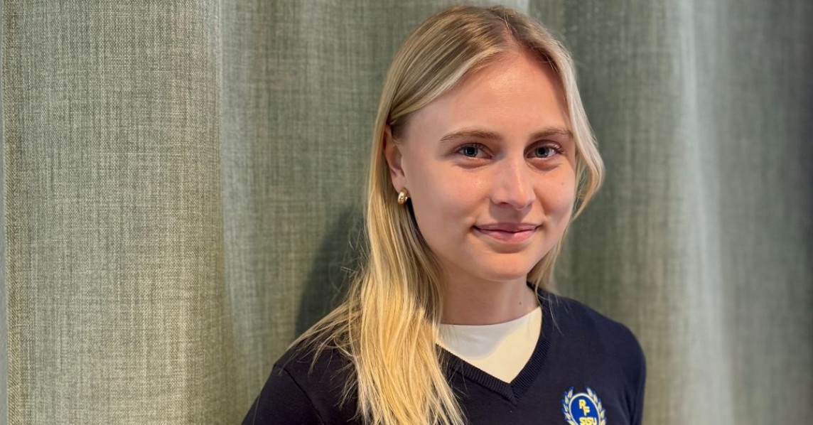 Inomhus, ljushårig, ung kvinna står mot en grågrön bakgrund. Hon småler in mot kameran. 